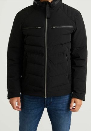 Homme portant une veste matelassée noire zippée avec des poches zippées poitrine et latérales, et un jean bleu, debout devant un fond uni.