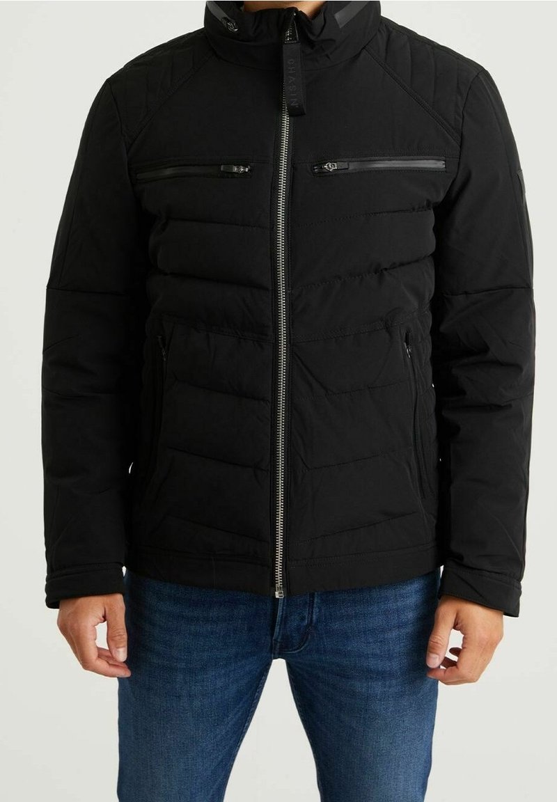 Mann trägt schwarze gesteppte Jacke mit Reißverschluss und Brust- sowie Seitenreißverschlusstaschen und blaue Jeans, steht vor einfachem Hintergrund.