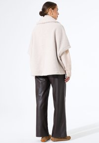 Beige strukturierte, übergroße Fleecejacke mit breitem Kragen, kombiniert mit dunkelbraunen Lederhosen und hellbraunen Schuhen, von hinten betrachtet.