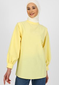 Haut à manches longues jaune avec un col montant et des poignets froncés, fabriqué dans un tissu lisse. Assorti avec un jean en denim. Le modèle pose de manière décontractée.