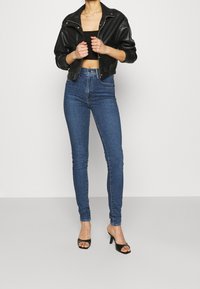 Chaqueta de cuero negra recortada, top ajustado negro sin mangas, vaqueros skinny de tiro alto en azul y sandalias negras de tacón.