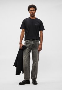 Μαύρη βαμβακερή μπλούζα με κοντά μανίκια, συνδυασμένη με ξεθωριασμένα γκρι παντελόνια με ίσια γραμμή. Το μοντέλο κρατά μια σκούρα ζακέτα. Μαύρα παπούτσια ολοκληρώνουν την εμφάνιση.