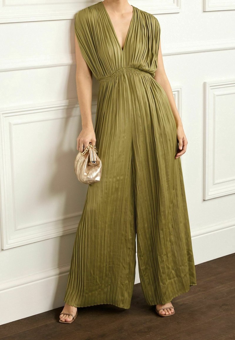 Femme portant une combinaison pantalon plissée vert olive à jambes larges avec un décolleté en V profond, tenant un sac à main doré métallique, debout contre un mur blanc à panneaux.