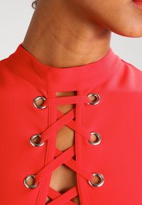 Närbild av nacke och bröst som bär ett livligt rött plagg med silverringar och korsade läderband vid framkant av halsringningen.