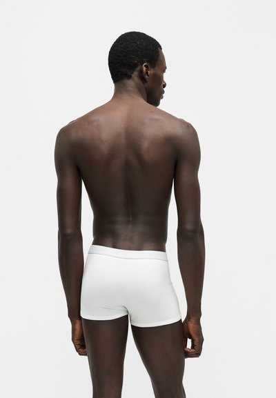 Sort mand stående med ryggen til iført hvide boxershorts mod en ensartet lys grå baggrund.