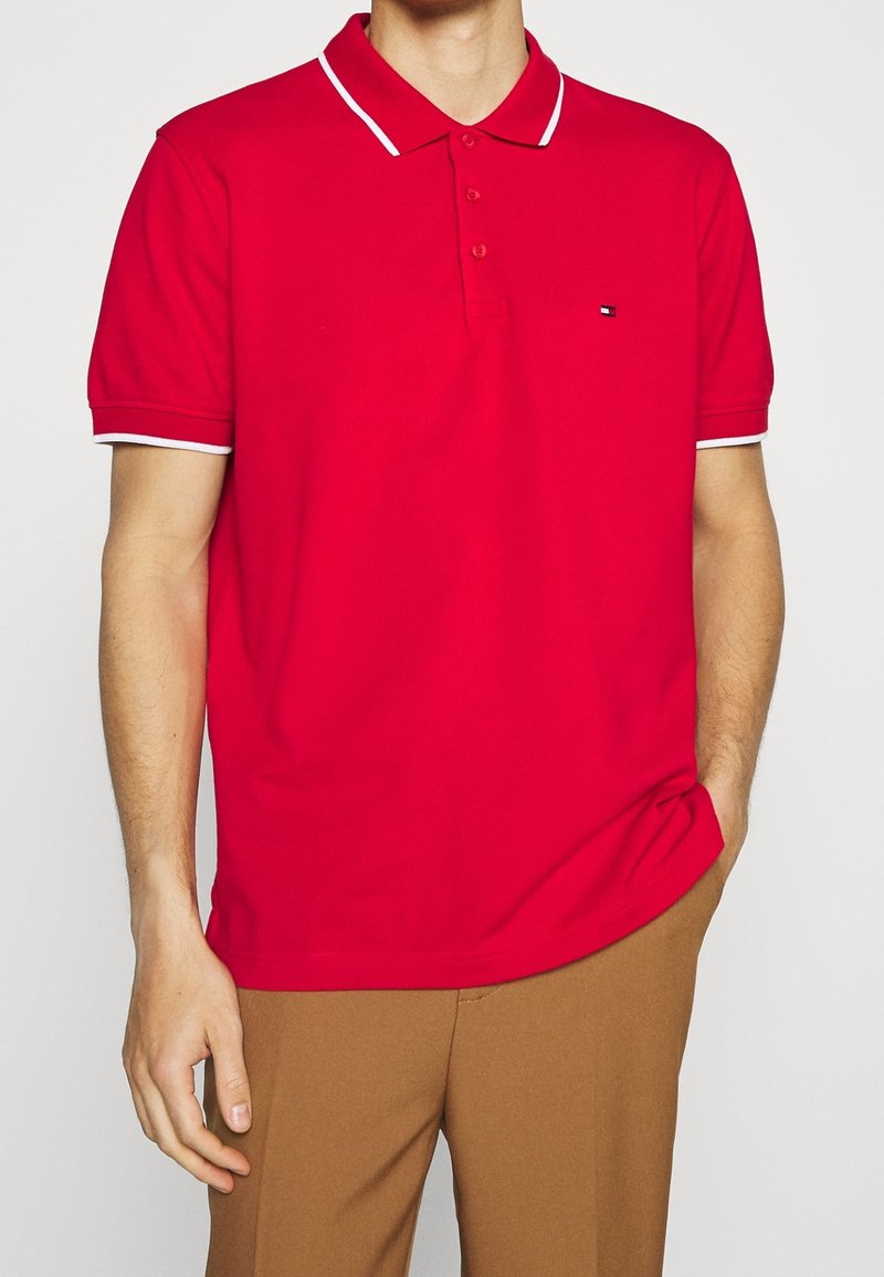 Polo rouge au design classique, avec une patte à trois boutons et un liseré blanc sur le col et les manches. Tissu texturé et petit détail de logo.