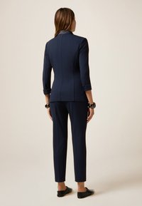 Marineblauer maßgeschneiderter Anzug mit tailliertem Blazer und schmal zulaufenden Hosen. Glattes Material, Dreiviertelärmel und keine sichtbaren Verschlüsse. Schwarze flache Schuhe.