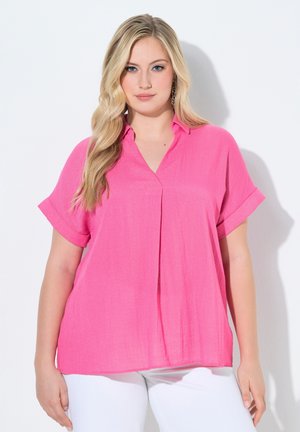 Blonde vrouw met een felroze blouse met korte mouwen en een witte broek, staand tegen een effen lichte achtergrond.