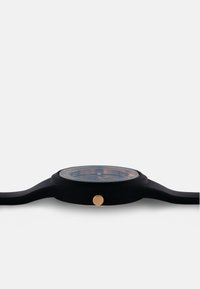 Orologio in silicone nero con cassa rotonda, quadrante trasparente, dettagli dorati e un pulsante laterale. Cinturino piatto con una texture liscia.