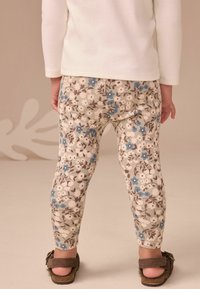Bloemenlegging met een geribbelde textuur, met blauwe en bruine bloemen op een crèmekleurige achtergrond. Draagt met bruine sandalen.
