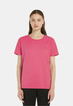 Giovane donna con capelli castani lunghi, che indossa una T-shirt rosa semplice a maniche corte e pantaloni neri, rivolta in avanti su uno sfondo bianco.