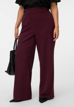 Frau trägt hoch taillierte, weit geschnittene burgunderfarbene Hose, schwarzen Top, schwarze spitze Schuhe und trägt eine schwarze Handtasche in der linken Hand.