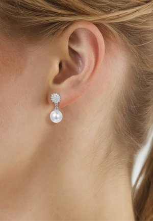 Close-up van het oor van een vrouw die een zilveren oorknopje draagt met een cluster van kleine steentjes en een hangende witte parel.