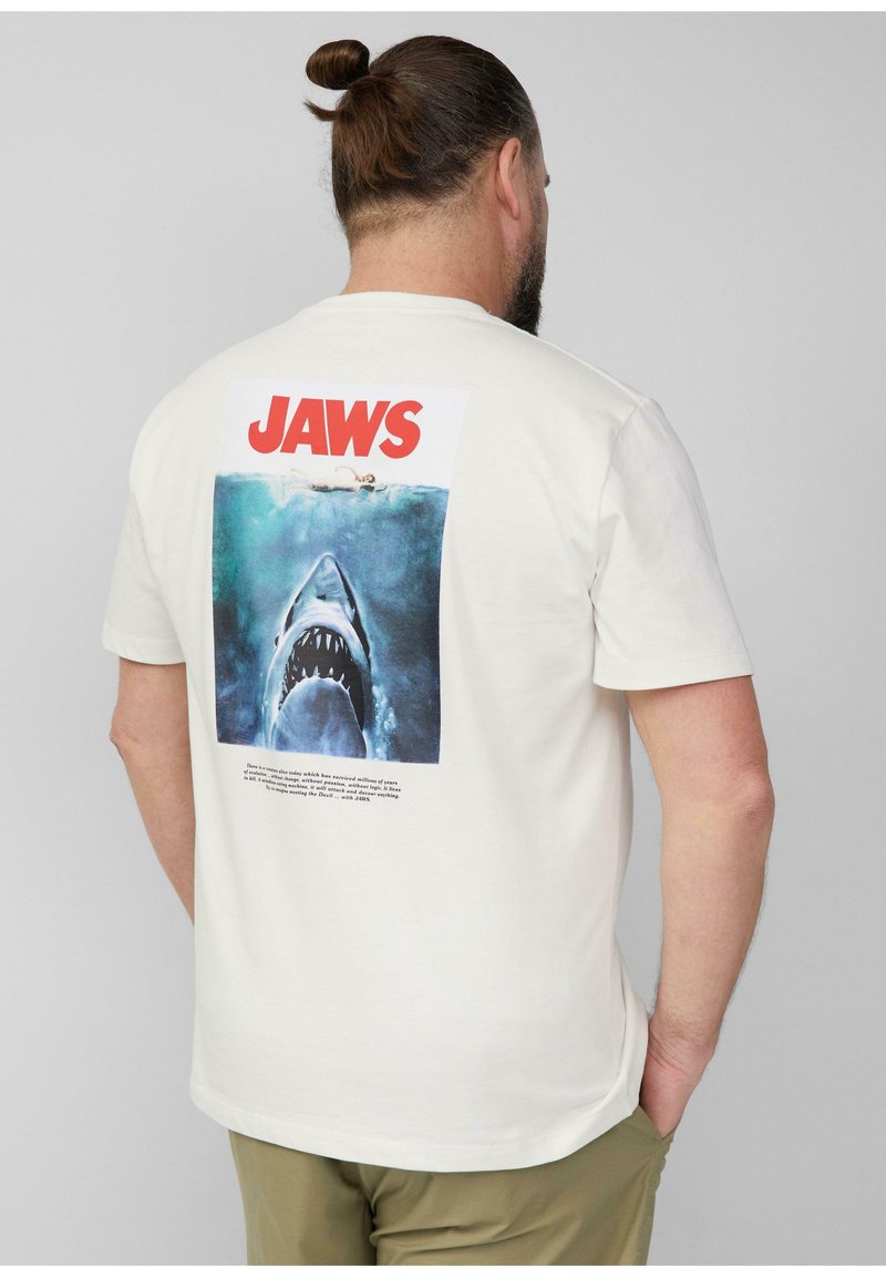 Crème-kleurig katoenen t-shirt met een grote afbeelding van een haai uit "Jaws" op de achterkant, vergezeld van vetrode tekst en een bijbehorende beschrijving eronder.