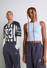 Dvaja dospelí, oblečení v športovom oblečení Adidas; jeden má na sebe čiernobiele pruhované tričko s dlhým rukávom, druhý má svetlomodrý crop top a fialové nohavice.