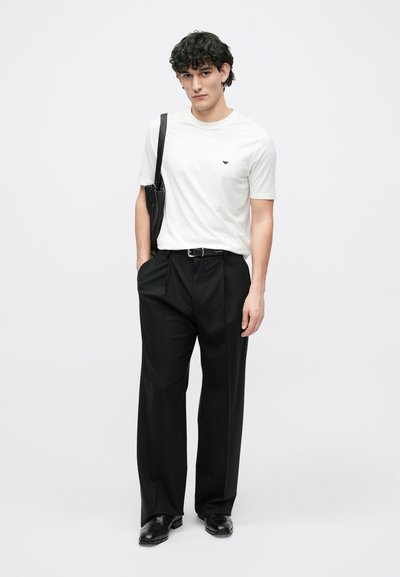 Fehér rövid ujjú póló kis fekete logóval, fekete pliszírozott nadrággal és fekete cipővel párosítva; a modell egy fekete táskát tart.