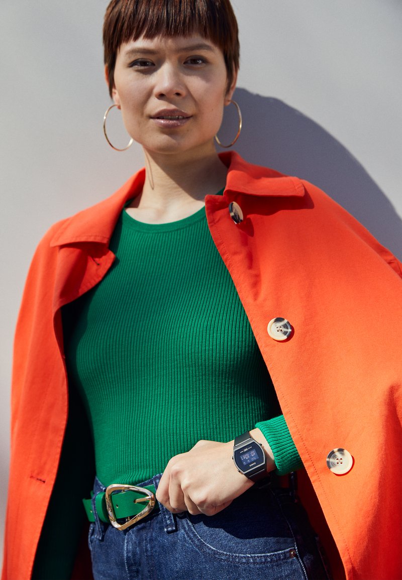 Montre numérique noire, haut à côtes vertes, veste orange, jeans bleus, ceinture à boucle dorée, boucles d'oreilles dorées, avec des détails de boutons sur la veste.