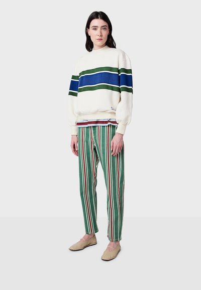 Krémová mikina so zelenými a modrými pruhmi, spárovaná so zelenými pruhovanými nohavicami s červenými a bielymi akcentami. Neutrálne zásuvné topánky.