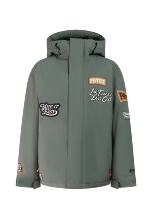 Junge Person trägt eine graue Mütze, grüne Winterjacke mit Aufnähern, schwarze Handschuhe, lockere schwarze Hose und schwarze Schuhe, steht vor einem schlichten Hintergrund.