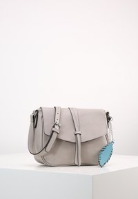 Světle šedá kožená taška s klopou, nastavitelným popruhem a připevněným modrým koženým lístkem jako charm. Hladký povrch, minimalistický design.