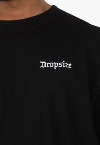 Fekete pamut póló fehér hímzett "Dropsize" felirattal. Klasszikus kerek nyakkivágással, rövid ujjakkal és sima textúrával.