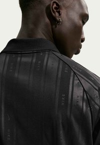 Κοντινό πλάνο ενός ατόμου που φοράει μαύρο μπλουζάκι Nike με διακριτικές κάθετες ρίγες και λογότυπα, στραμμένο πλευρικά, με ένα ασημένιο σκουλαρίκι κρίκο στο αυτί.