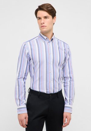 Giovane uomo che indossa una camicia a maniche lunghe, abbottonata, con strisce verticali blu, bianche e rosa, abbinata a pantaloni neri, in piedi davanti a uno sfondo bianco.