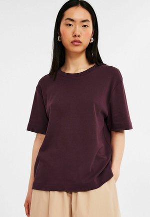 Short-sleeve, loose-fitting t-shirt in deep purple cotton. Features a round neckline and soft texture, paired with beige pants.