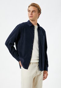 Camisa azul marino de botones con cuello amplio, mangas largas y un bolsillo en el pecho, combinada con una camiseta de cuello redondo de color claro y pantalones beiges.