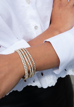 Fyra guldtonade armband prydda med klara kristaller, med en smal och elegant design. Bäraren har armen vilande mot en vit skjorta med knappar.