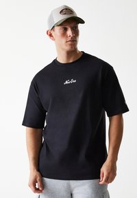 Black short-sleeve t-shirt with a crew neck, featuring white "New Era" logo on the chest. Model wears light grey sweatpants and a cap.