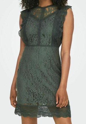 Hihaton tummanvihreä pitsimekko, jossa on kukkakuosi, aaltoilevat reunat ja läpikuultava kaula-aukko, pukeutuneena kiharatukkainen henkilö.