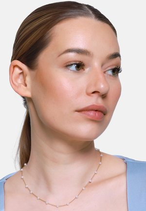 Junge Frau mit heller Haut und braunem, zurückgebundenem Haar, die eine zarte Goldkette mit gleichmäßig verteilten kleinen Perlen und ein hellblaues Oberteil trägt.