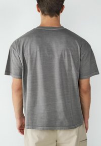 Szara koszulka z krótkim rękawem o luźnym kroju, gładkiej fakturze i okrągłym dekolcie, z minimalnymi detalami szycia wzdłuż krawędzi.