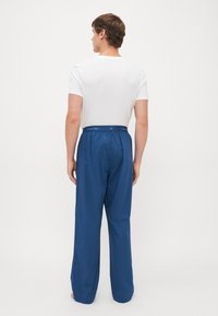 Muškarac u bijeloj majici s kratkim rukavima i plavim Calvin Klein hlačama stoji bos okrenut leđima na plain bijeloj pozadini.