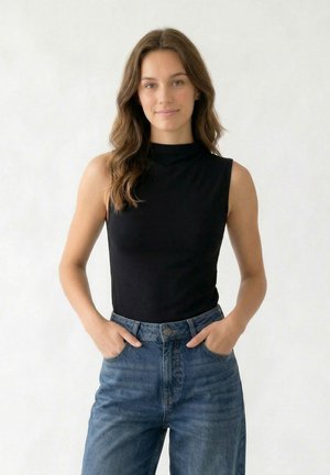 Noor naine pikkade laineliste juustega, kandes varrukateta musta topi ja kõrge vöökohaga siniseid teksapükse, käed taskutes, ühtlase taustaga.