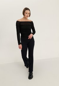 Černý top s odhalenými rameny má shromážděnou texturu a je spárován s tmavými proužkovanými kalhotami. Model má na sobě černé mohutné boty. Neutrální pozadí.