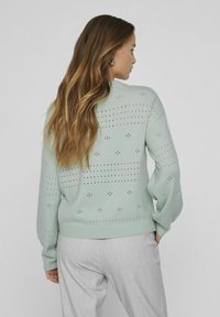 Světle zelený pletený svetr s otvorovými vzory a žebrovanými manžetami. Stylizováno s světle šedými kalhotami, které zdůrazňují uvolněný střih.