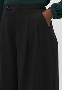 Zwarte broek met een getailleerde pasvorm, plooien en zijzakken. Gemaakt van een gladde stof met een klassieke knoopsluiting in de taille.