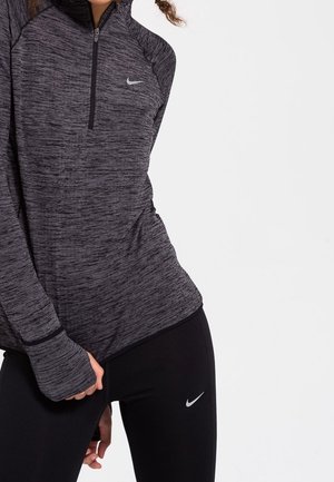 Woman wearing a grey long-sleeve athletic zip-up top and black leggings with Nike logos, shown from mid-torso to mid-thigh.