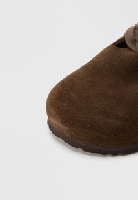 Καφέ καστόρινο παπούτσι με στρογγυλεμένη μύτη, υφή στην επιφάνεια και λεπτή, ευέλικτη ελαστική σόλα. Ο σχεδιασμός περιλαμβάνει π λαϊνές ανοίγματα για αναπνοή.