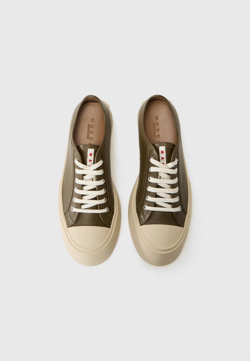 Une paire de baskets basses vert olive avec des semelles épaisses beiges et des lacets blancs, photographiée de dessus sur un fond blanc.