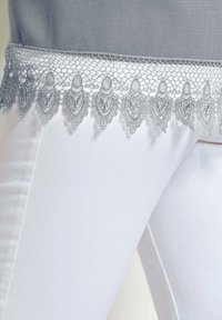 Weiße Hose mit grauem Spitzenbesatz, der einen gewellten Rand und filigrane Muster aufweist, wodurch eine zarte Textur vor einem glatten Hintergrund zur Geltung kommt.