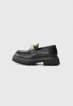 Mocasines de cuero negro con una suela de goma gruesa, que presentan un acento dorado en el empeine y un diseño de punta redondeada.