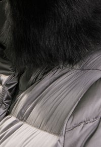 Černý kožešinový límec na šedé prošívané bundě, který kombinuje hladké a prošívané části s lesklým povrchem, zvyšujíce teplo a texturu.