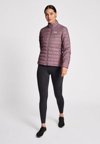 Lilla pufferjakke med stående krave, vandret quiltning og logo på brystet. Båret med sorte leggings og sorte atletiske sko.