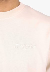 Hellrosa Rundhals-T-Shirt aus weichem Stoff, mit einem eingestickten "Dropsike"-Logo in Weiß auf der Brust.