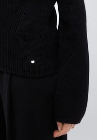 Pull noir en laine avec un motif texturé, des manches larges et un petit logo circulaire sur l'ourlet. Tissu lisse et épais.