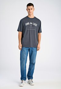 Camiseta gris de manga corta con "JUNK de LUXE" impreso en blanco, combinada con jeans azul de pierna recta y zapatillas blancas con acentos coloridos.