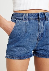 Džínové šortky s vysokým pasem, obsahující boční kapsy, ohrnutý lem a viditelné švy v kontrastní nitě. Barva je středně modrá.